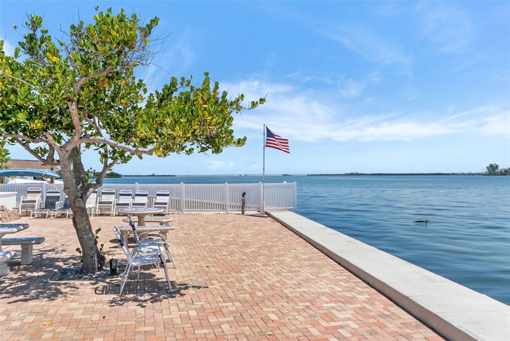 A paved patio with a tree and seating overlooks a bay with a flag — 611 GULF DRIVE N UNIT C27, BRADENTON BEACH, FL