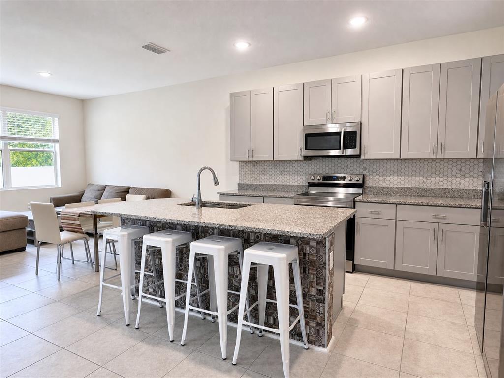 A kitchen features a granite-topped island with white stools, gray cabinetry, and stainless appliances — 1622 WAY OF PEACE LANE, MASCOTTE, FL
