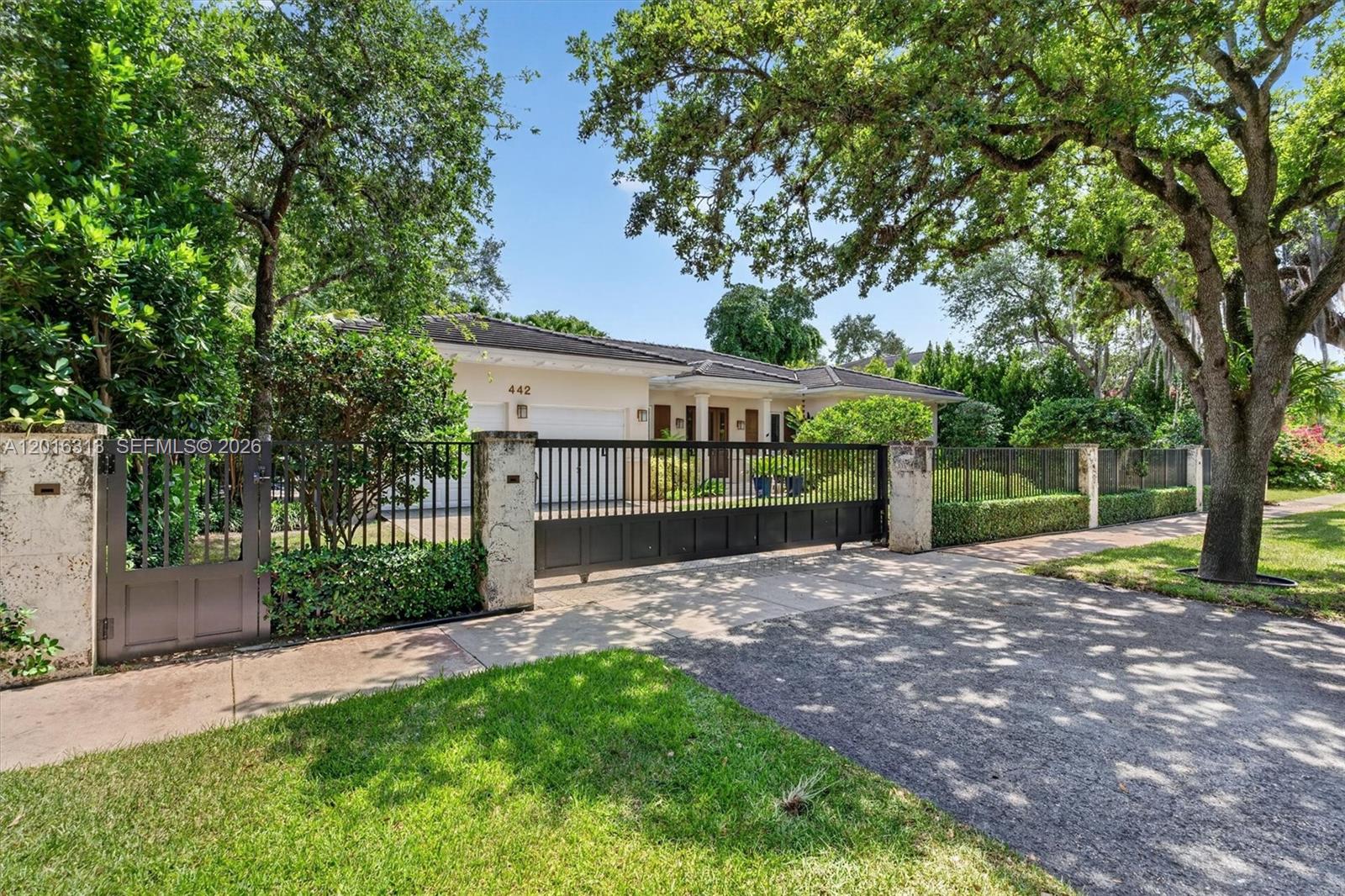 A gated driveway leads to a single-story house with a white exterior and mature trees — 442 Amalfi Ave, Coral Gables FL 33146, CORAL GABLES, FL