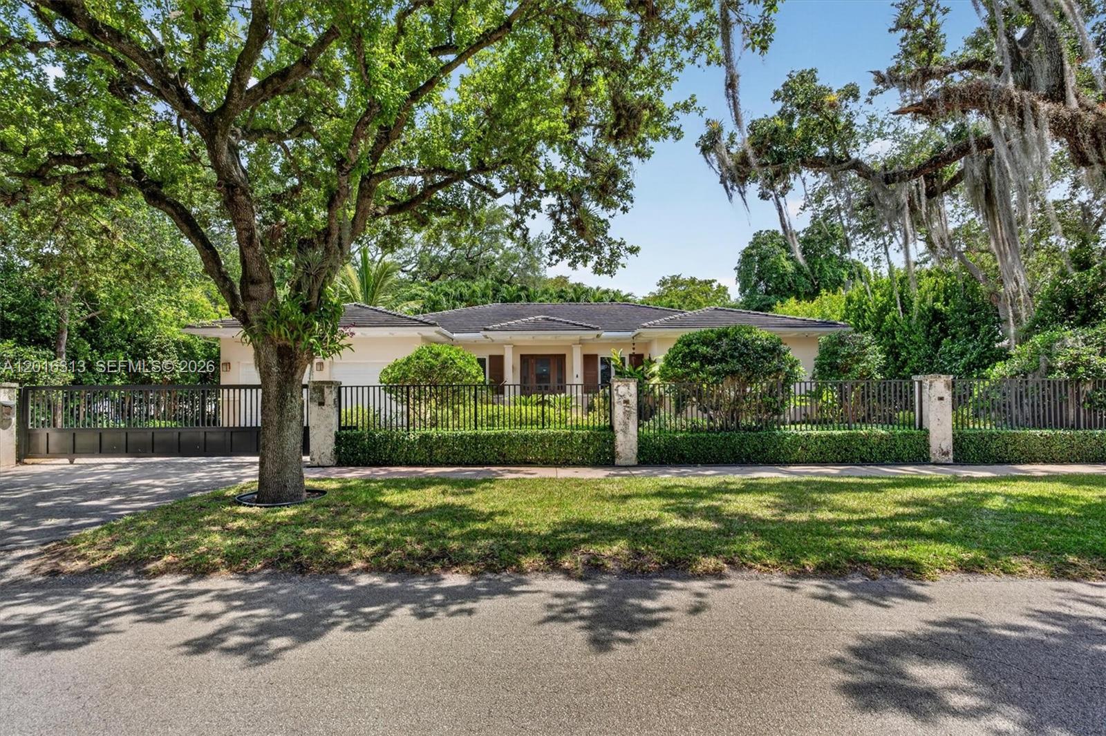 A single-story house with a dark roof sits behind a black metal fence and trees — 442 Amalfi Ave, Coral Gables FL 33146, CORAL GABLES, FL