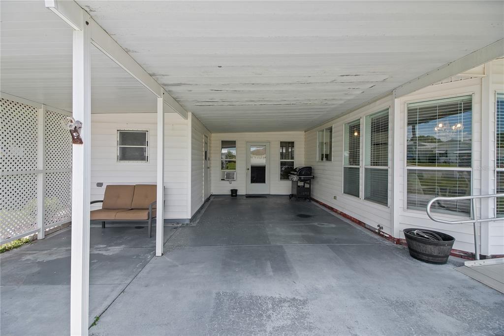 A covered carport with a concrete floor, white siding, and a small outdoor seating area — 1203 GRANADA COURT, THE VILLAGES, FL