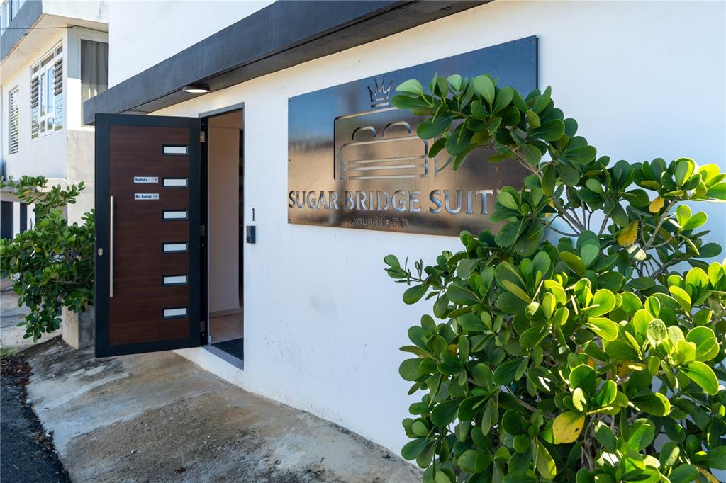 The exterior entrance of Sugar Bridge Suites features a modern door and metal signage — 2 BO. BORINQUEN, AGUADILLA, PR