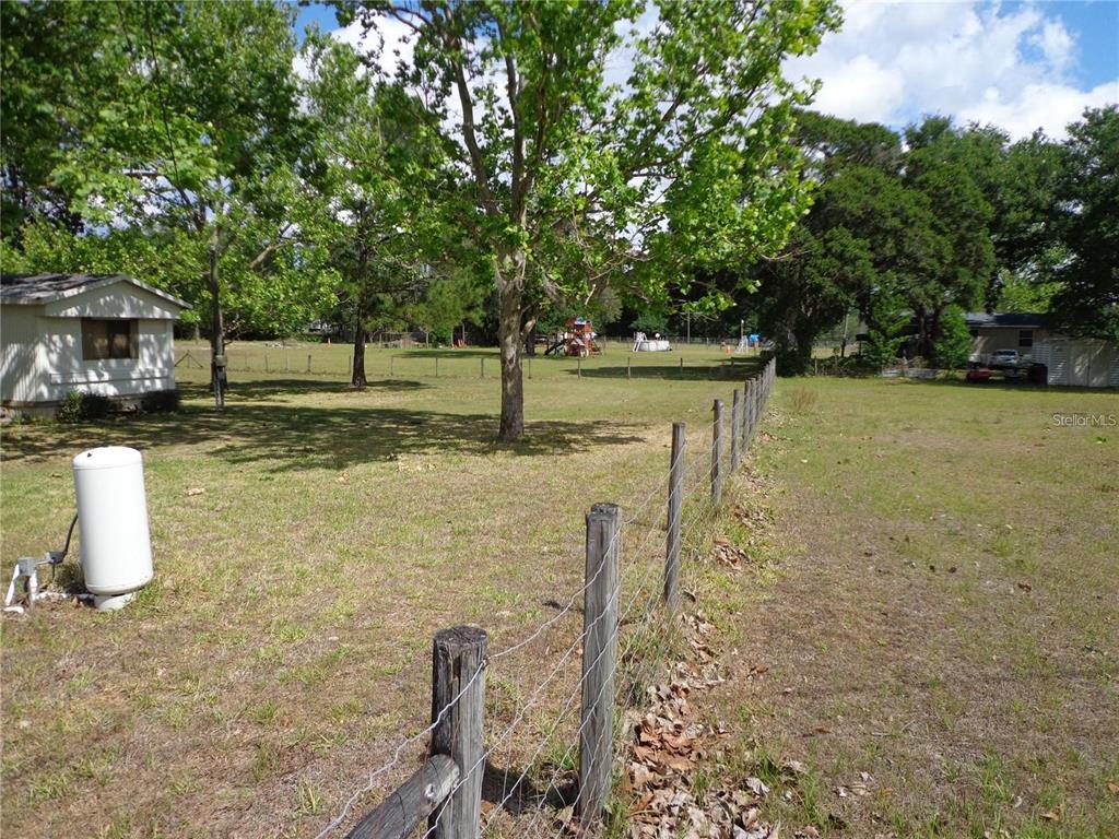 A fenced grassy yard features a large tree, a propane tank, and distant residential structures — 17911 ORANGE DRIVE, SPRING HILL, FL