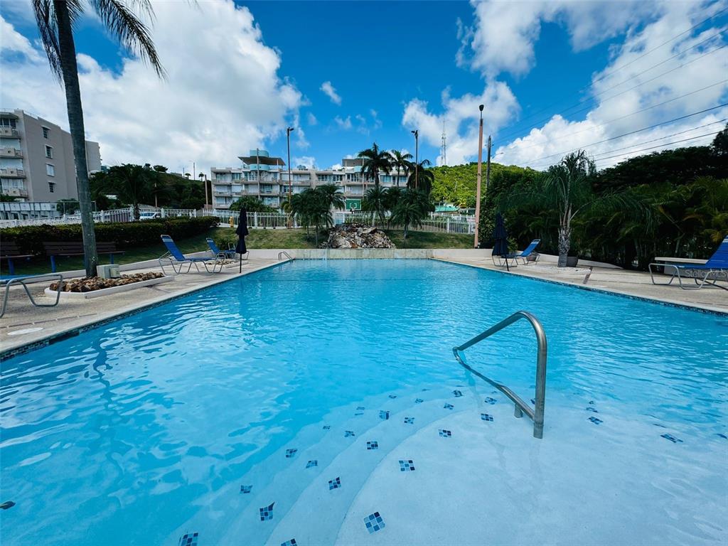An outdoor swimming pool with steps and lounge chairs surrounded by palm trees and buildings — COND. VILLAS DEL FARO 5015 PUNTA TUNA ST. UNIT 406, MAUNABO, PR