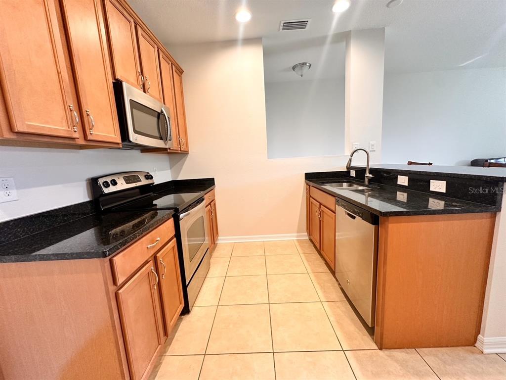 A kitchen features light wood cabinets, black countertops, stainless steel appliances, and beige tile flooring — 11511 84TH STREET CIRCLE E UNIT 105, PARRISH, FL