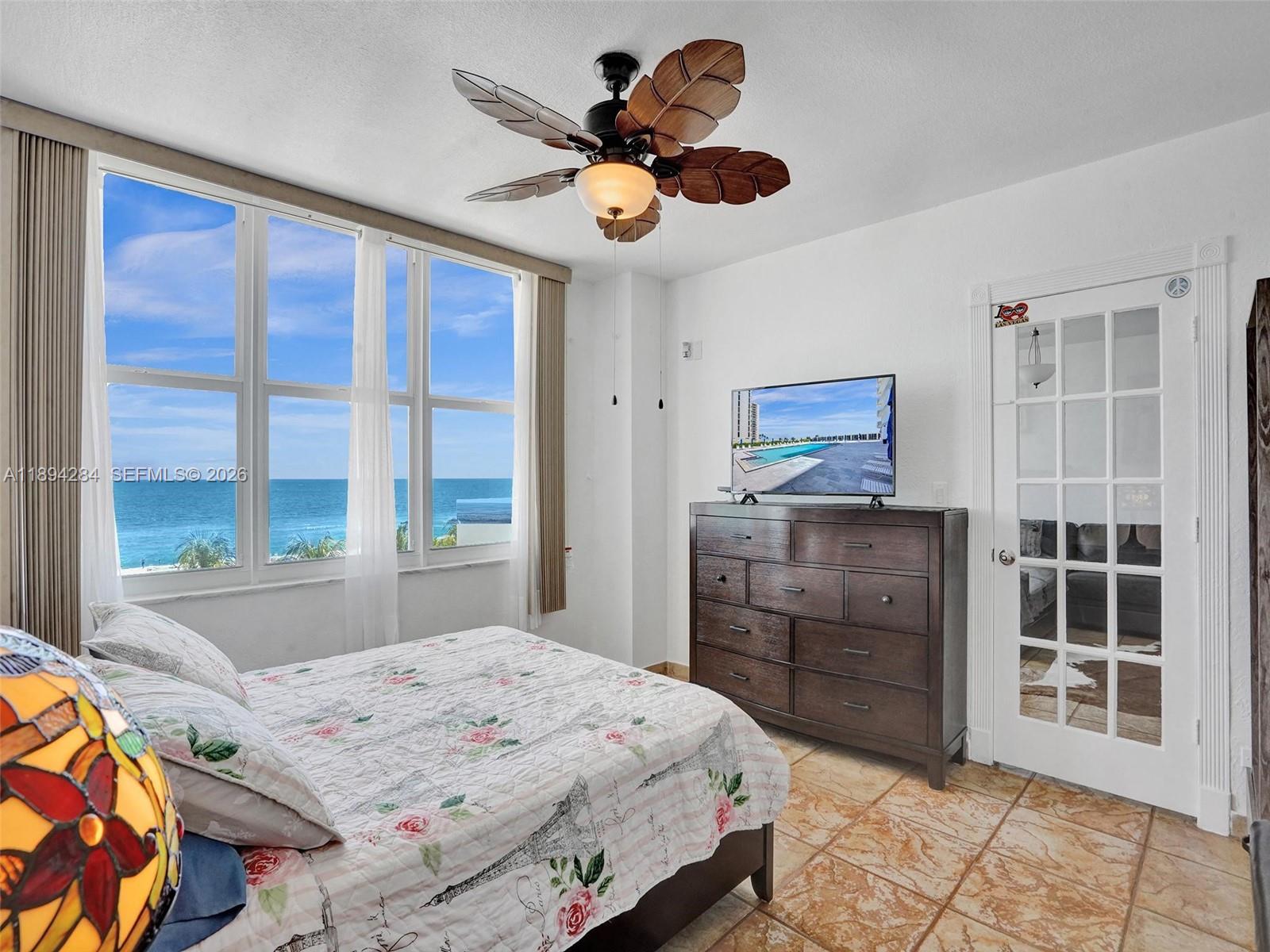 A bedroom features a floral bedspread, a dark wood dresser with a television, and ocean views — 5601 Collins Ave # 504, Miami Beach FL 33140, MIAMI BEACH, FL