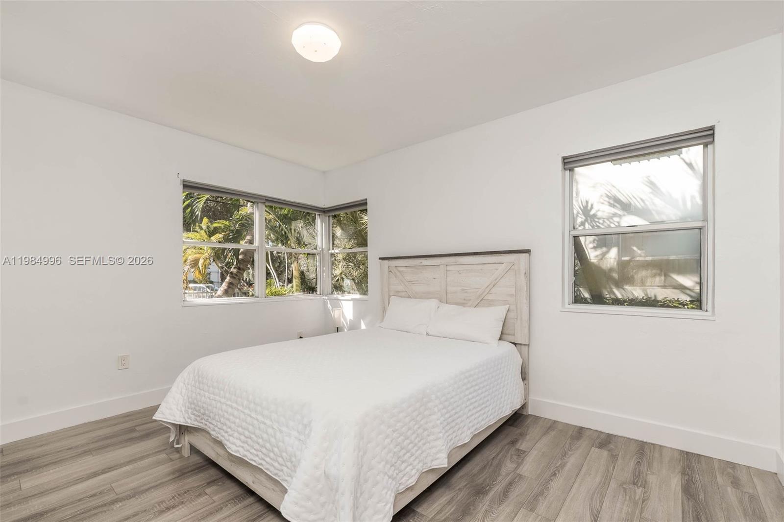 A bedroom features a white bed, light wood-look flooring, and windows with exterior palm trees — 1275 Marseille Dr # 35, Miami Beach FL 33141, MIAMI BEACH, FL