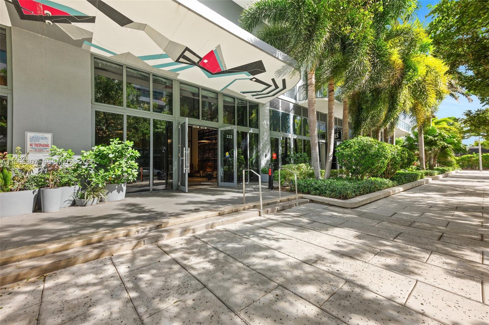 The exterior entrance of a modern building features a geometric mural, glass doors, and landscaping — 121 NE 34th St # 2314, Miami FL 33137, MIAMI, FL