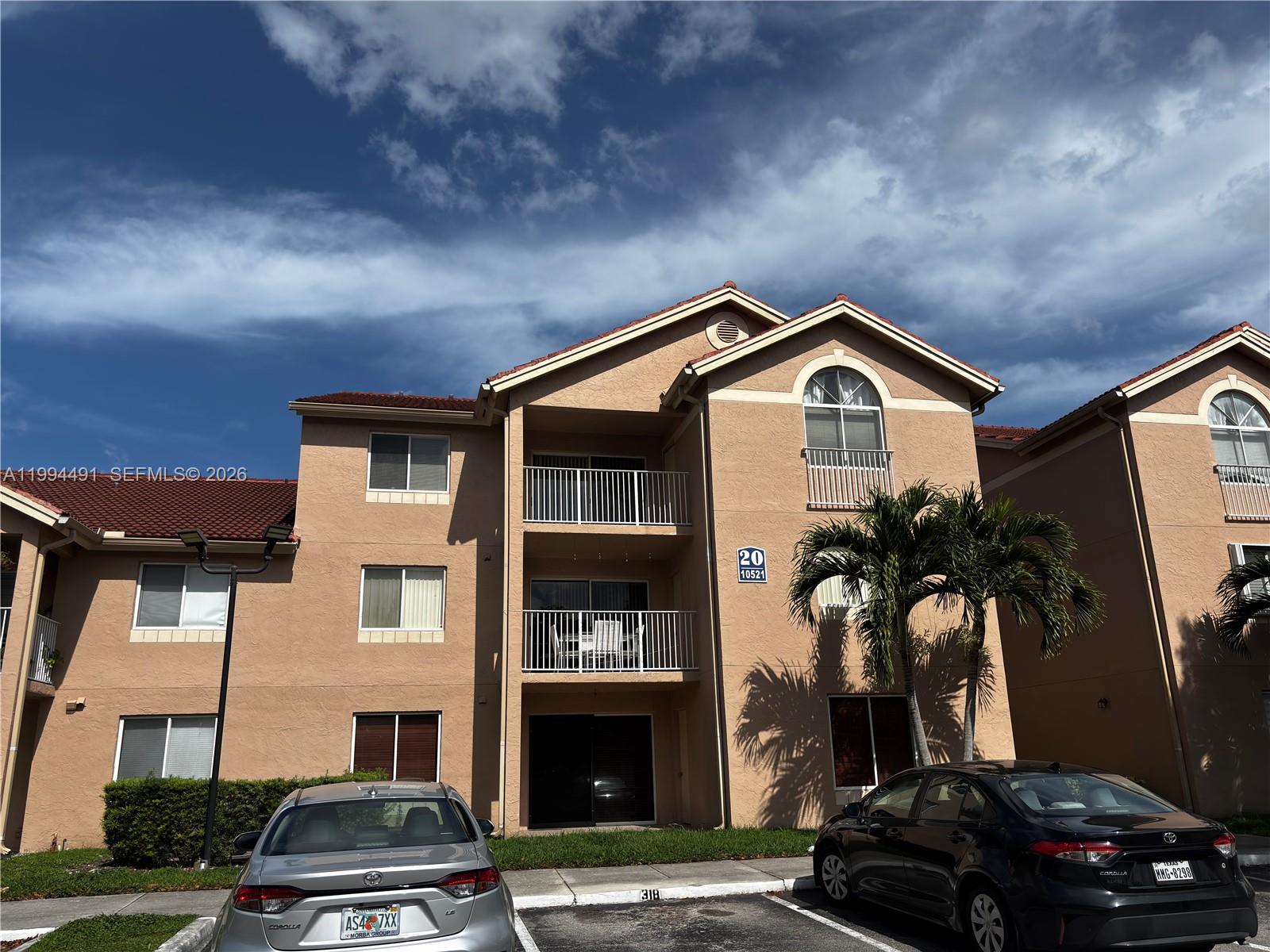 A three-story tan apartment building features balconies, a tiled roof, and a parking lot — 10521 SW 158th Ct # 303, Miami FL 33196, MIAMI, FL
