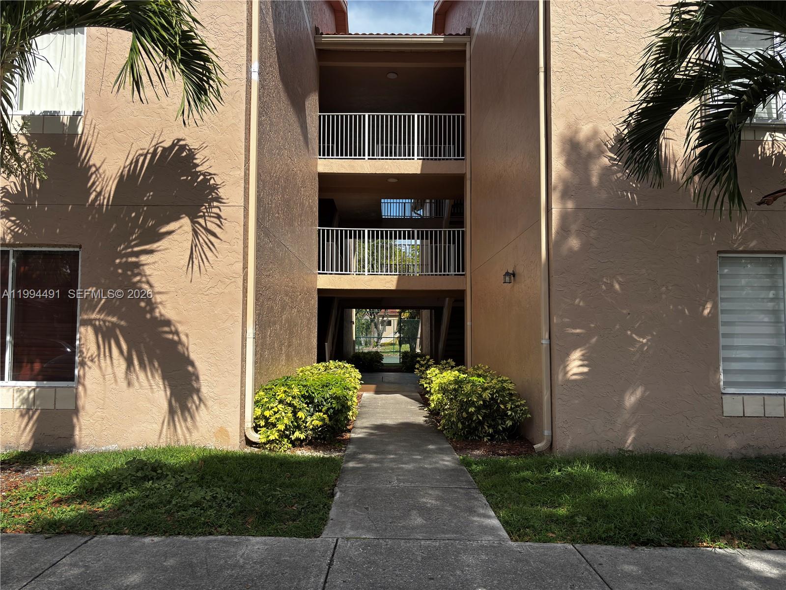 A concrete walkway leads between two tan apartment buildings with balconies and palm trees — 10521 SW 158th Ct # 303, Miami FL 33196, MIAMI, FL