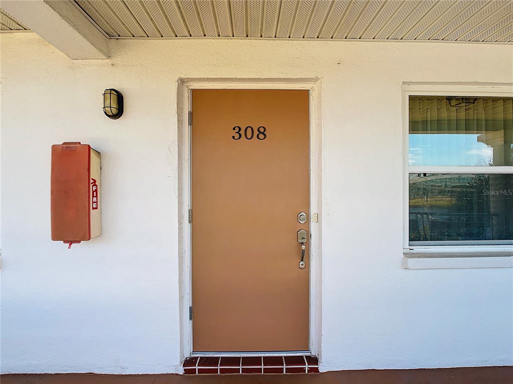 A brown entry door labeled 308 is set in a white exterior wall with a fire extinguisher — 6015 19TH STREET N UNIT 308, ST PETERSBURG, FL