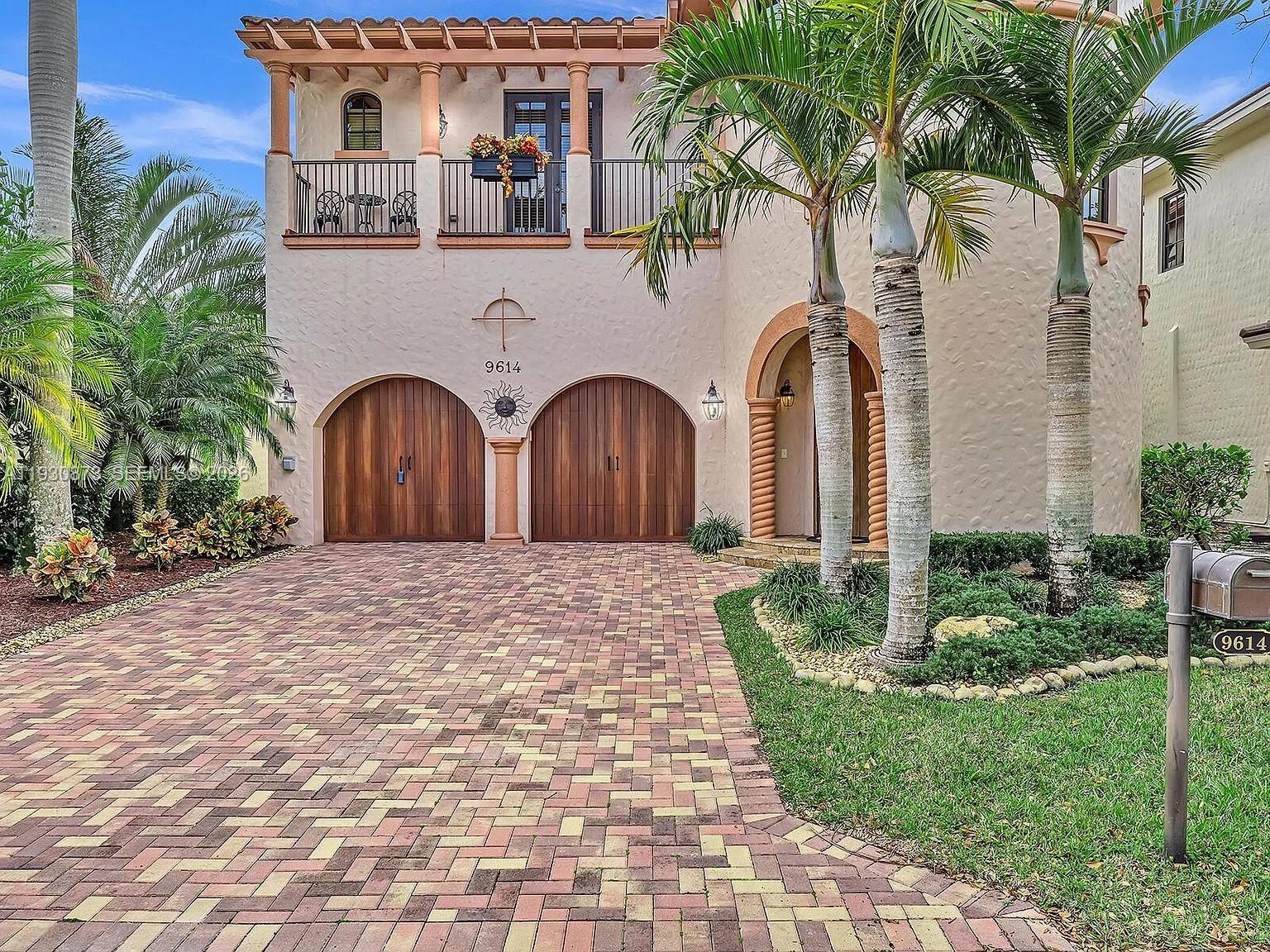 A two-story stucco house features arched wooden garage doors, a balcony, and a brick driveway — 9614 Kenley Ct, Parkland FL 33076, PARKLAND, FL