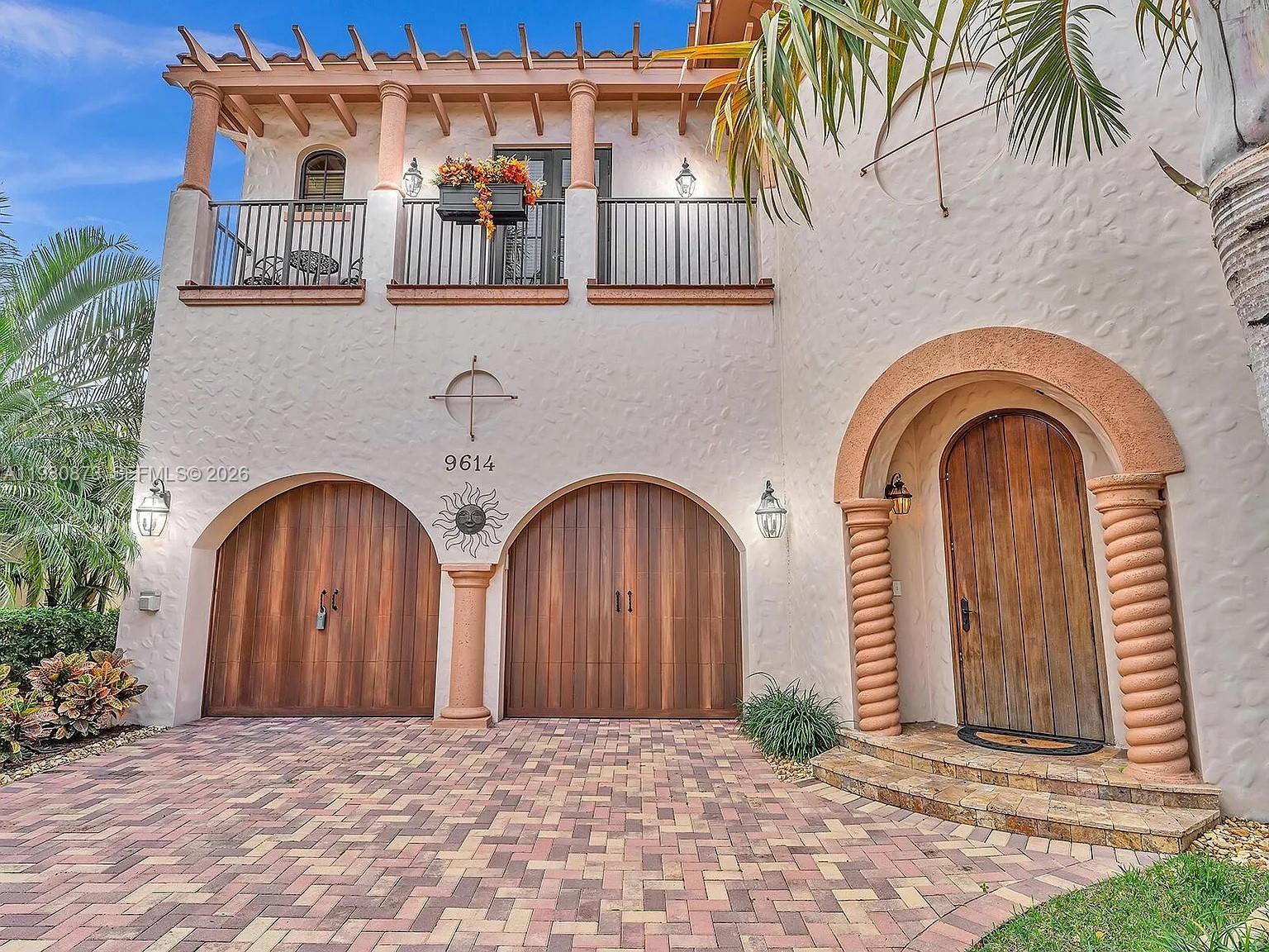 A two-story Mediterranean-style home features arched wooden garage doors, a stone entryway, and a balcony — 9614 Kenley Ct, Parkland FL 33076, PARKLAND, FL