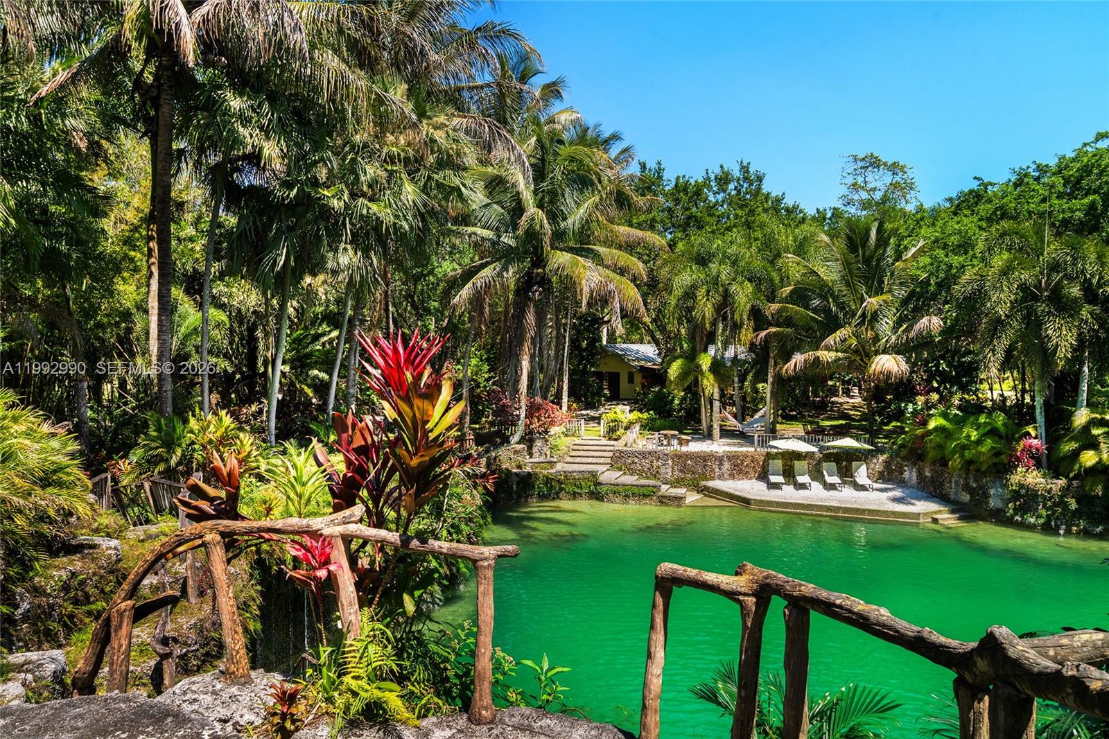 A lush tropical garden features a vibrant green swimming pond with a rustic wooden railing — 15000 SW 200th St, Miami FL 33187, MIAMI, FL