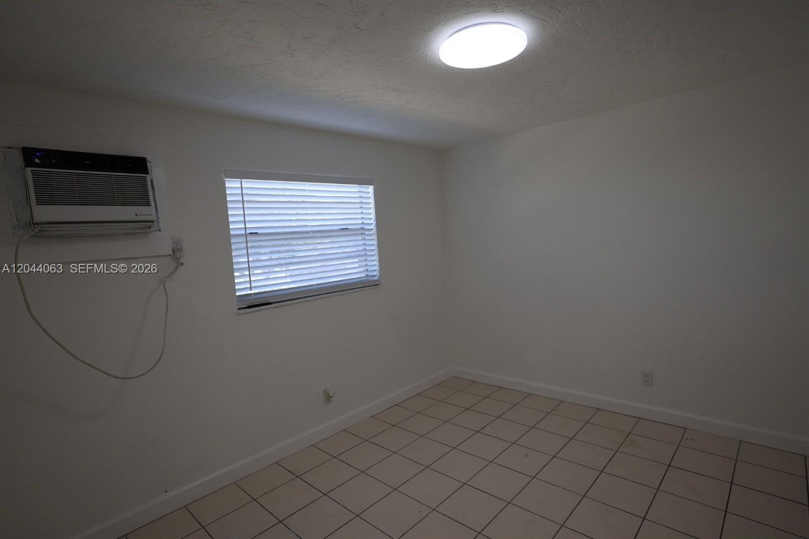 An empty room features white walls, tiled flooring, a window with blinds, and a wall-mounted air conditioner — 1235 W 66th St # 13, Hialeah FL 33012, HIALEAH, FL