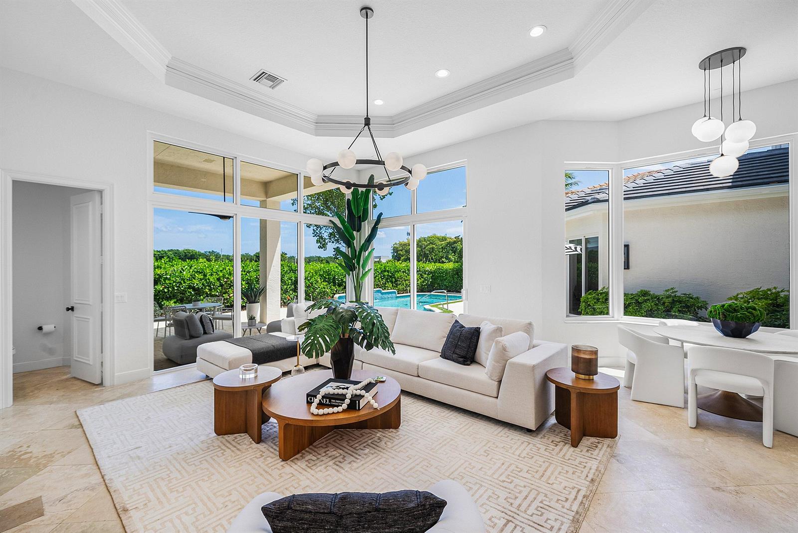 A bright living room features a white sofa, round wooden tables, and large windows overlooking a pool — 16032 Bristol Isle Way, Delray Beach FL 33446, DELRAY BEACH, FL
