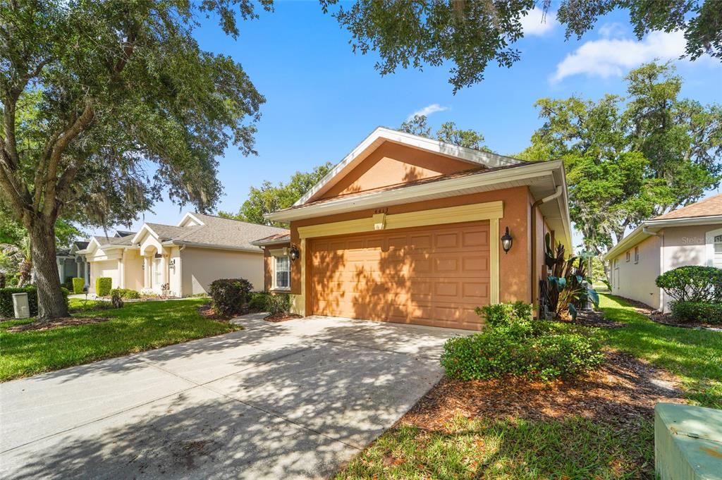 A tan single-story house with a two-car garage and a concrete driveway under large trees — 4413 CALIQUEN DRIVE, BROOKSVILLE, FL