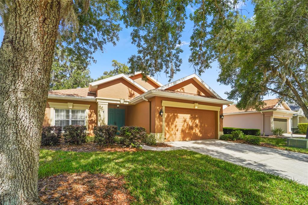 A single-story house with a tan stucco exterior, a two-car garage, and mature oak trees — 4413 CALIQUEN DRIVE, BROOKSVILLE, FL