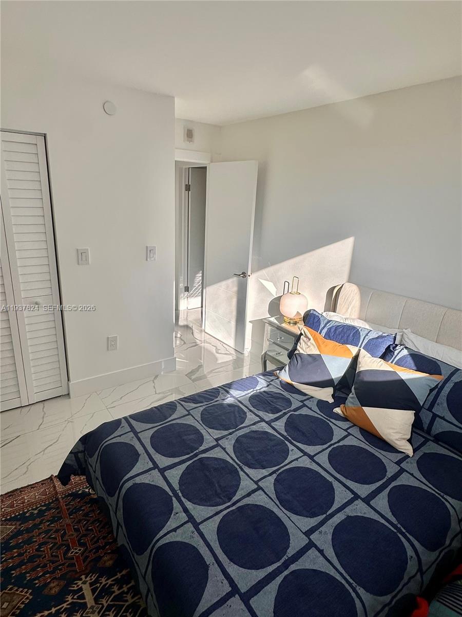A bedroom features a bed with a patterned blue quilt, white tile floors, and a doorway — 2457 Collins Ave # 408, Miami Beach FL 33140, MIAMI BEACH, FL