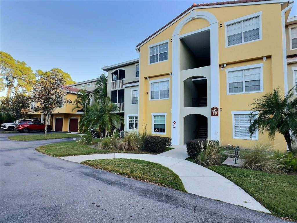 A three-story yellow apartment building with a central open-air stairwell and surrounding landscaping — 4751 TRAVINI CIRCLE UNIT 4-105, SARASOTA, FL