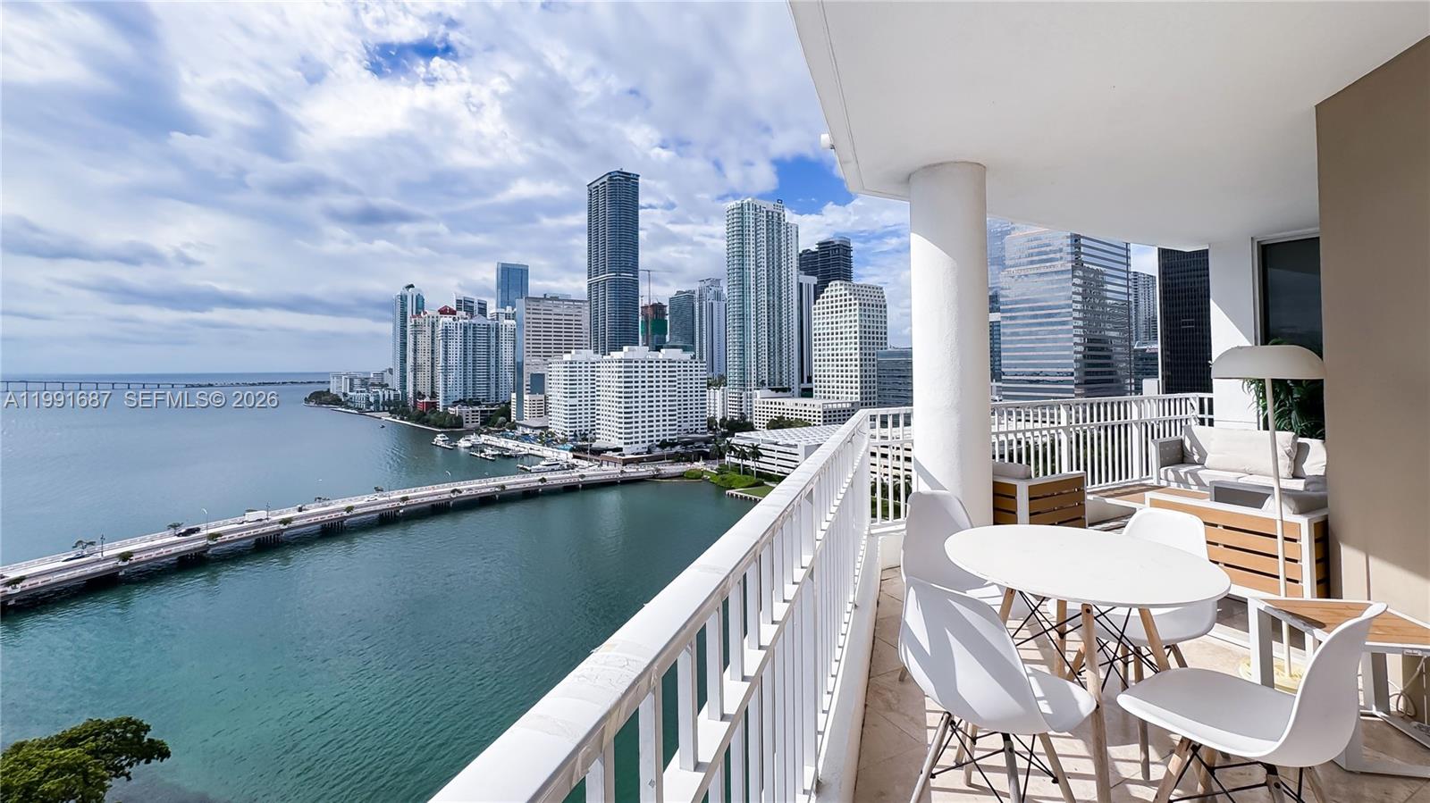 A balcony with white furniture overlooks a bay, a bridge, and a city skyline — 701 Brickell Key Blvd # 1912, Miami FL 33131, MIAMI, FL