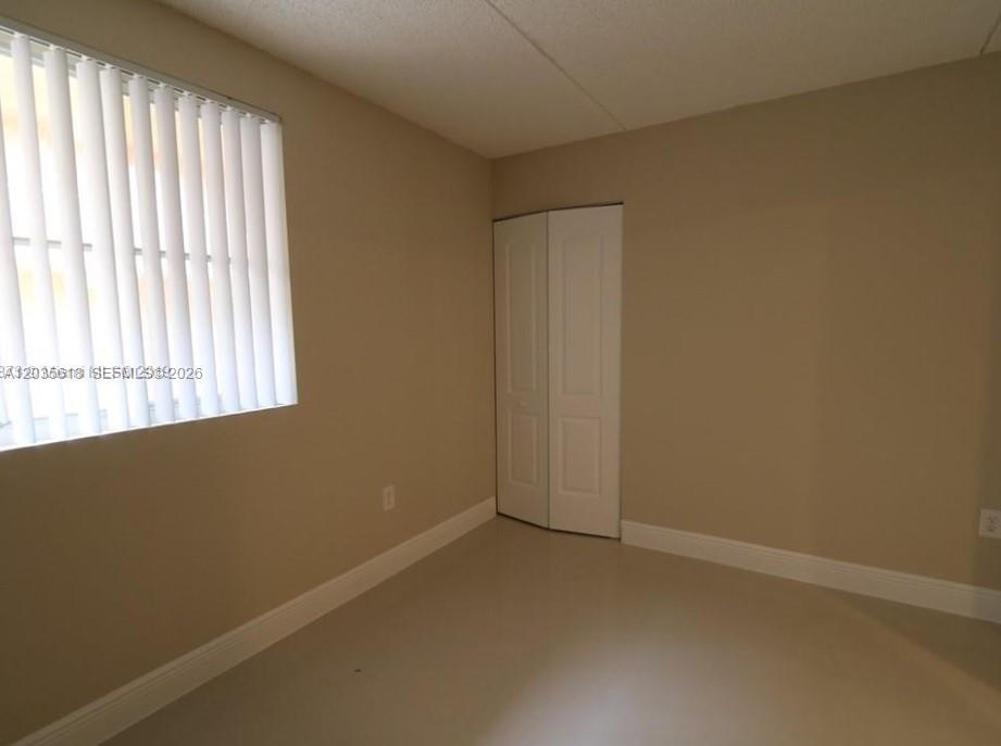 An empty bedroom features beige walls, light-colored flooring, a window with vertical blinds, and a closet — 6175 NW 186th St # 305, Hialeah FL 33015, HIALEAH, FL