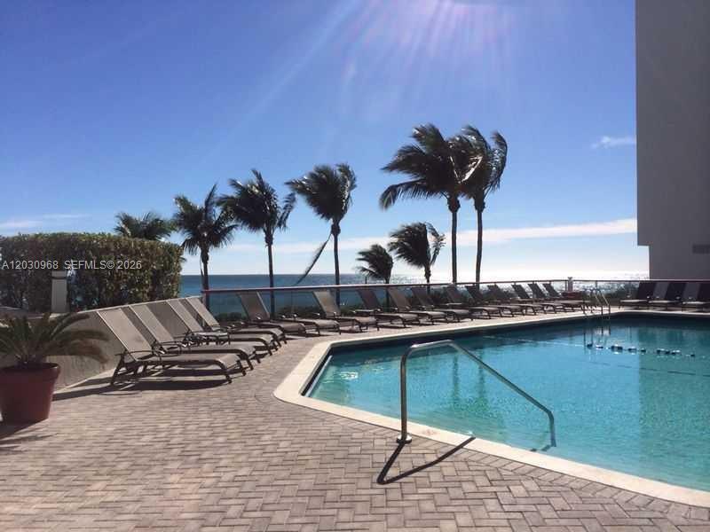 A swimming pool with lounge chairs on a brick patio overlooking the ocean and palms — 6767 Collins Ave # 206, Miami Beach FL 33141, MIAMI BEACH, FL