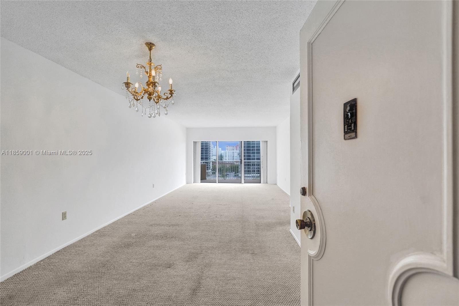 An empty living room features beige carpeting, a crystal chandelier, and a balcony with city views — 1965 S Ocean Dr # 7K, Hallandale Beach FL 33009, HALLANDALE BEACH, FL