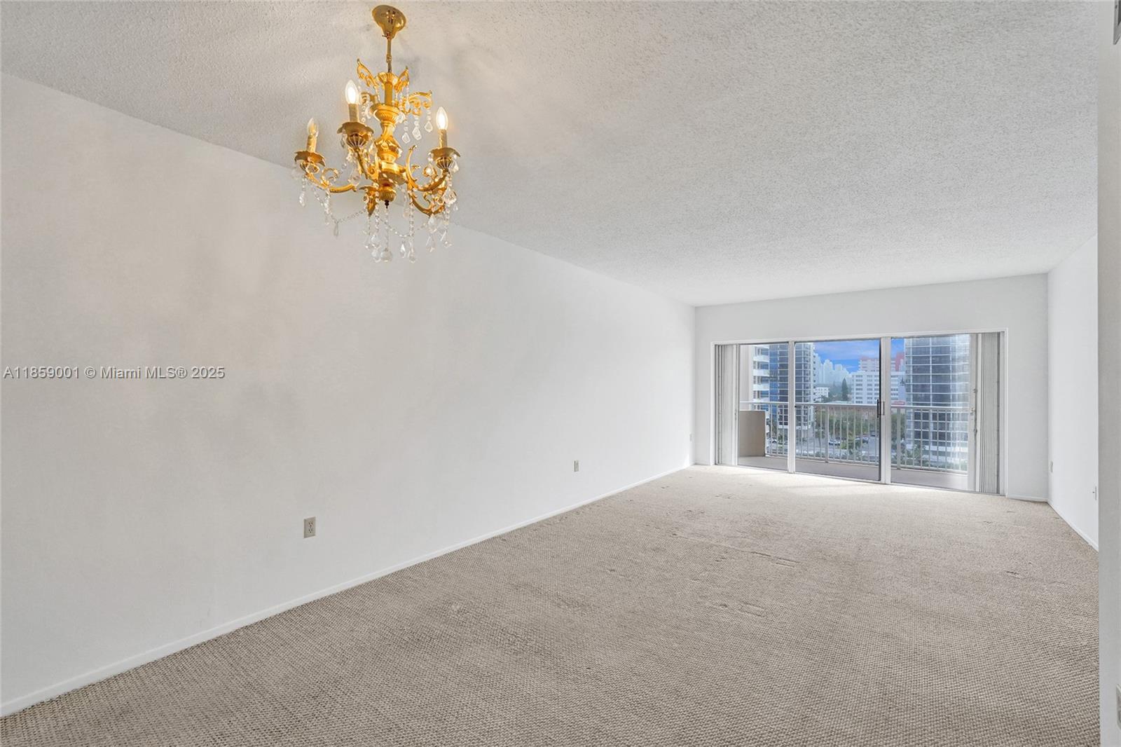 An empty room features beige carpeting, white walls, a gold chandelier, and a sliding glass door — 1965 S Ocean Dr # 7K, Hallandale Beach FL 33009, HALLANDALE BEACH, FL