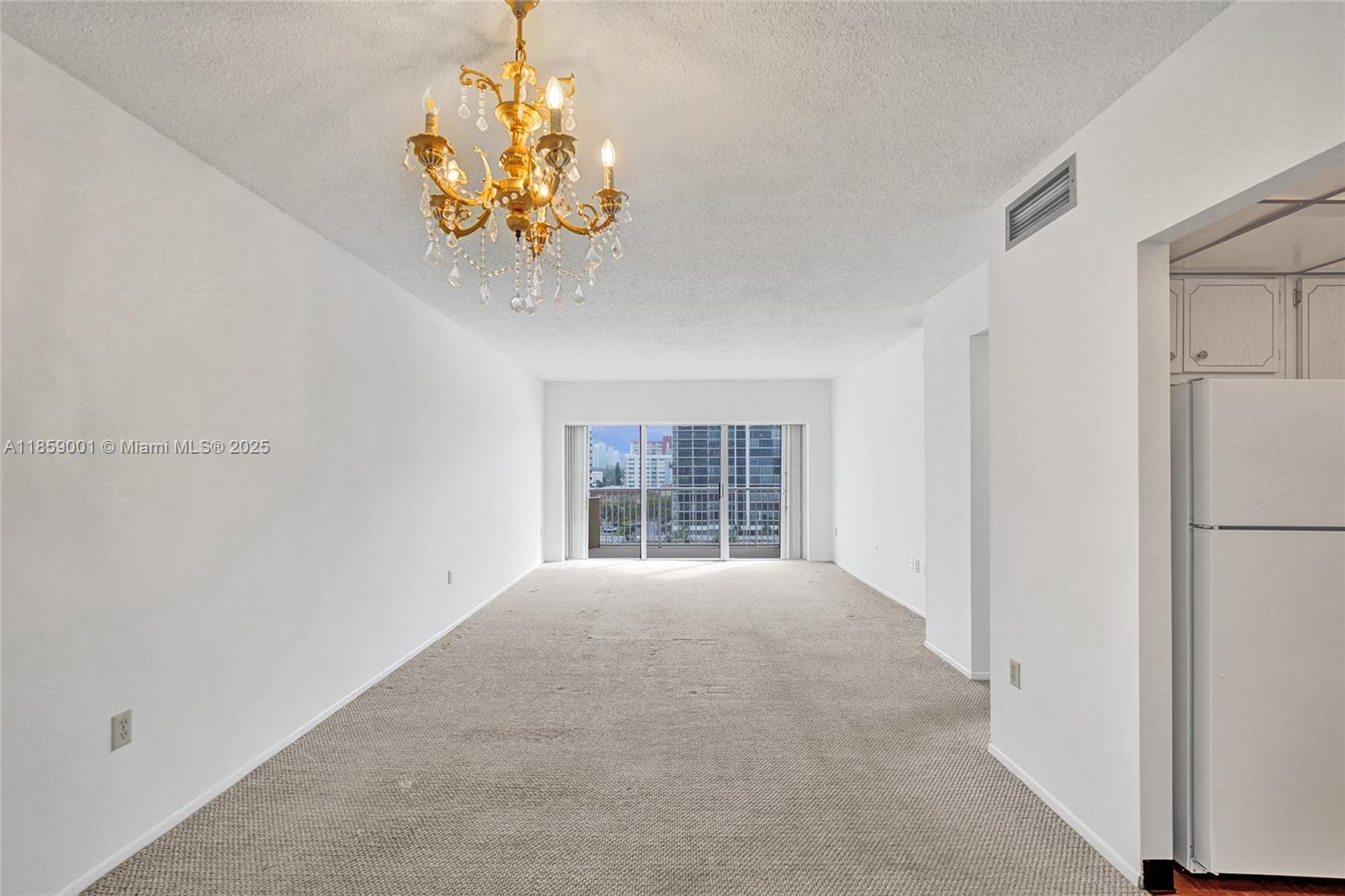 An empty living room features beige carpeting, white walls, a gold chandelier, and a balcony — 1965 S Ocean Dr # 7K, Hallandale Beach FL 33009, HALLANDALE BEACH, FL