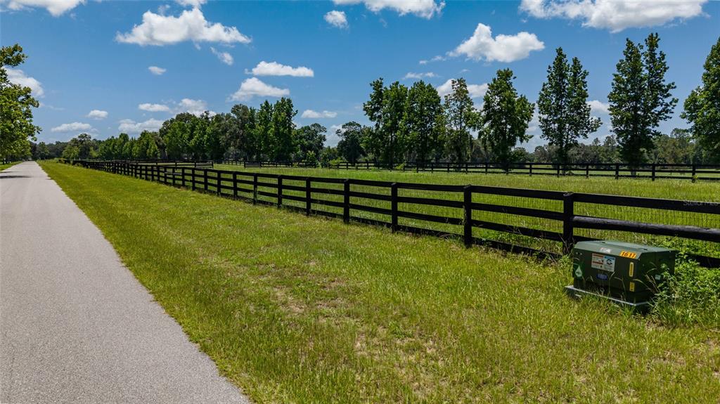 A paved road runs alongside a grassy field enclosed by a black wooden fence — SE 159TH LN., SUMMERFIELD, FL