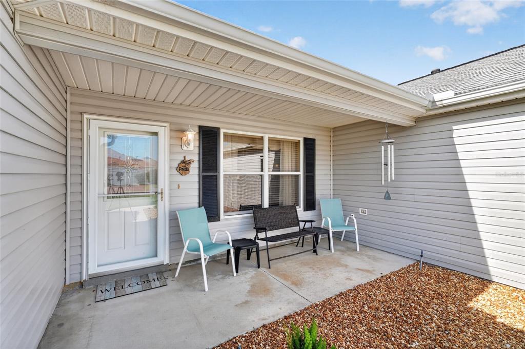 A covered concrete porch features light gray siding, a white door, and three outdoor chairs — 1952 ARDMORE WAY, THE VILLAGES, FL