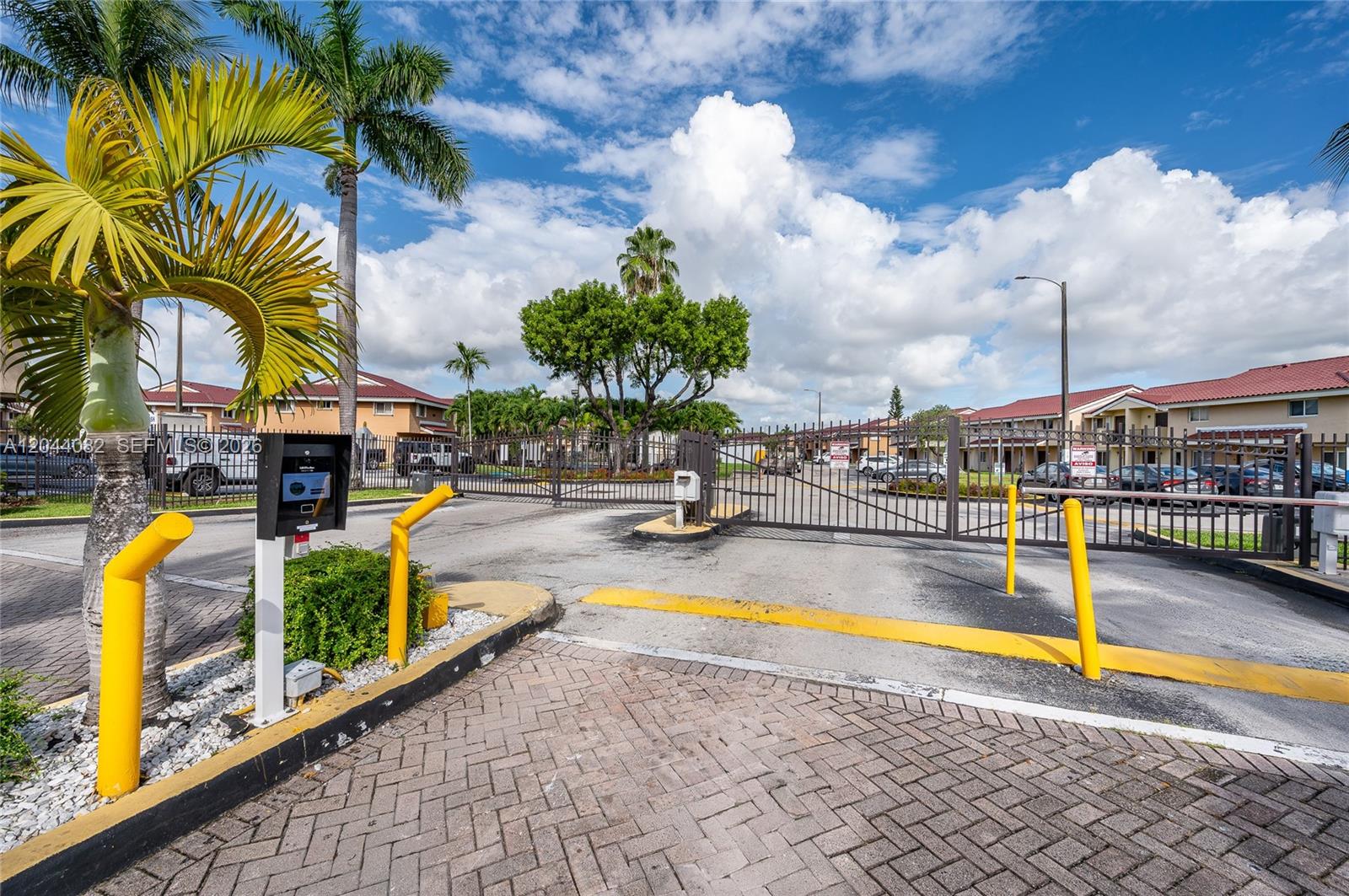 A gated entrance with a security keypad, yellow bollards, and palm trees under a cloudy sky — 550 NW 82nd Ct # 383, Miami FL 33126, MIAMI, FL