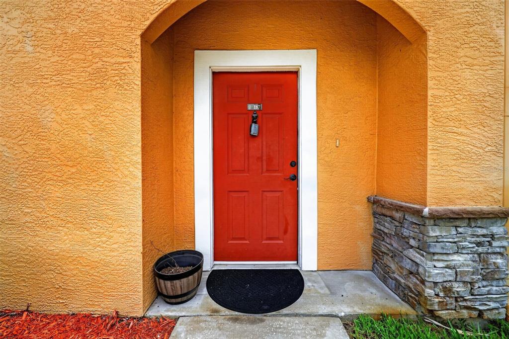 A red front door with a lockbox sits under an arched entryway with orange stucco — 2217 TUSCANY TRACE UNIT 7, PALM HARBOR, FL