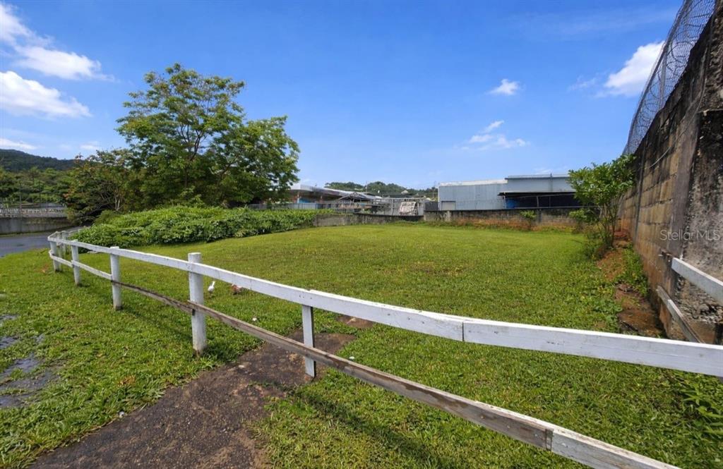 A grassy, fenced-in vacant lot sits next to a concrete wall under a blue sky — Carretera 1 BARRIO QUEBRADA ARENA, SAN JUAN, PR