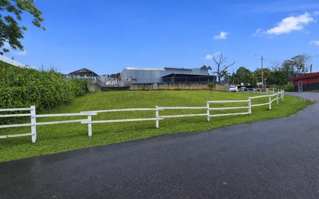 A grassy, sloped vacant lot is enclosed by a white fence along a paved road — Carretera 1 BARRIO QUEBRADA ARENA, SAN JUAN, PR