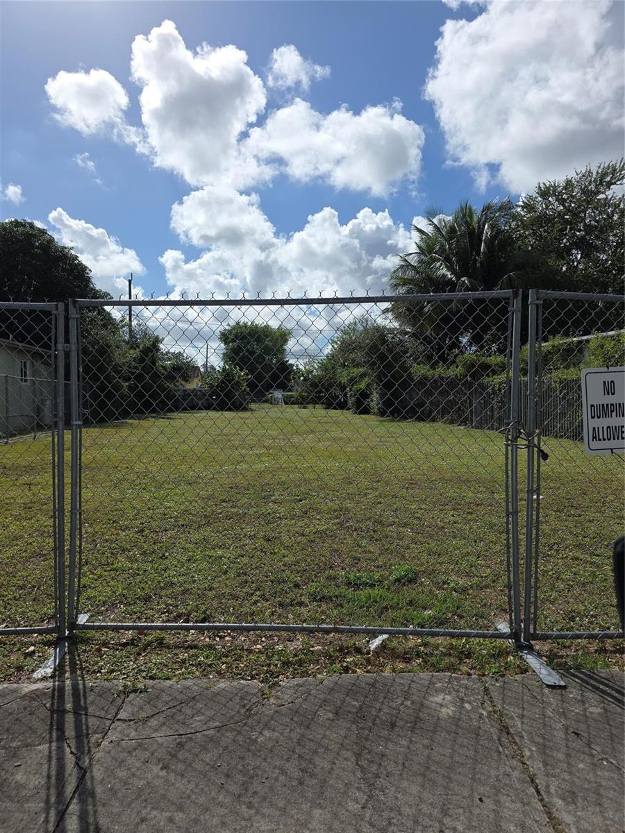 A fenced-in, vacant grassy lot is viewed from a concrete sidewalk under a cloudy sky — 1810 NW 84th Street, Miami FL 33147, MIAMI, FL