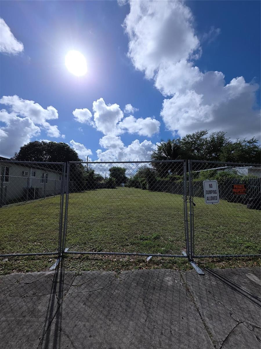 A fenced, vacant grassy lot is viewed from a concrete driveway under a sunny sky — 1810 NW 84th Street, Miami FL 33147, MIAMI, FL