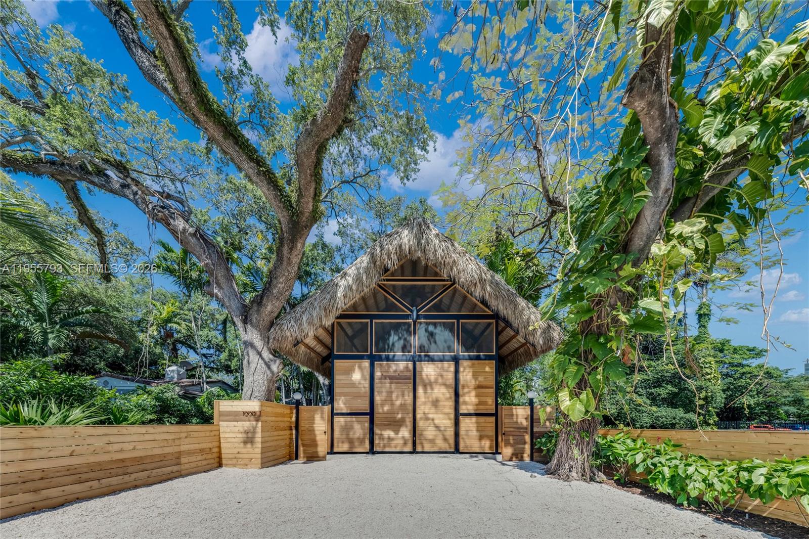 A thatched-roof structure with wooden doors stands behind a fence on a gravel driveway — 1000 NW 9th Ct, Miami FL 33136, MIAMI, FL