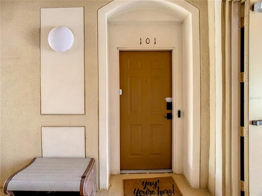 A tan front door marked 101 features a welcome mat and an adjacent wall light — 7664 COMROW STREET UNIT 101, KISSIMMEE, FL