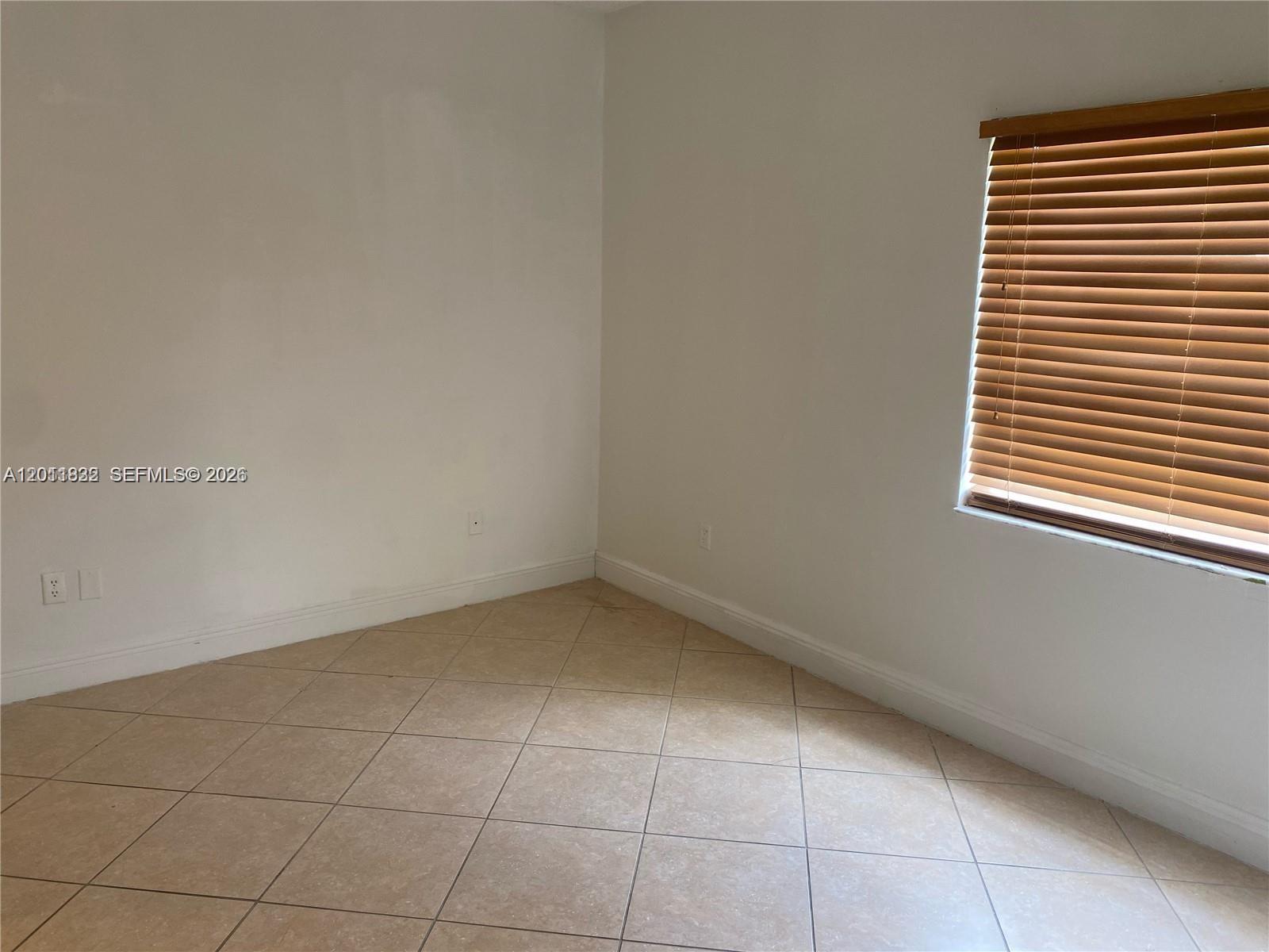 An empty room features white walls, beige tile flooring, and a window with wooden blinds — 18330 NW 68th Ave # G, Hialeah FL 33015, HIALEAH, FL