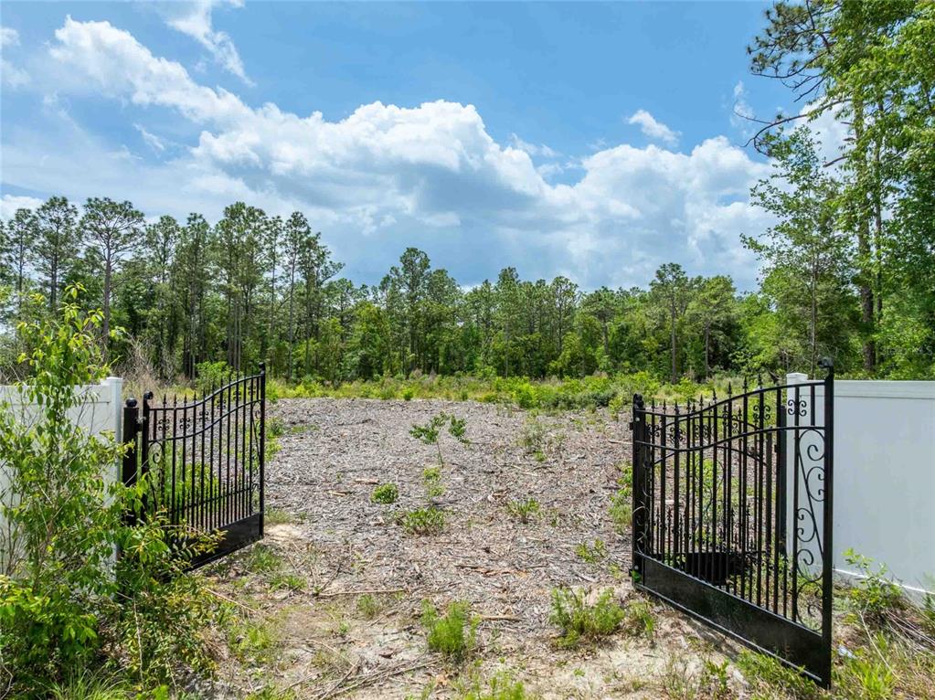 An open black metal gate leads to a cleared, wooded lot under a cloudy sky — 15022 GAR STREET, SPRING HILL, FL