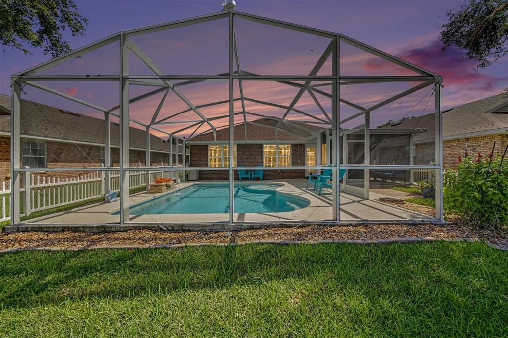 A swimming pool enclosed by a screened lanai sits behind a brick house at sunset — 567 CORAL TRACE BOULEVARD, EDGEWATER, FL
