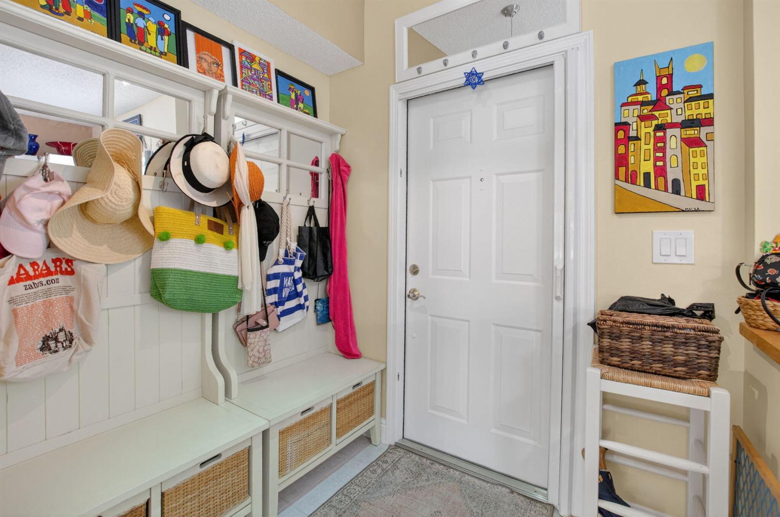 An entryway features a white storage bench with wicker baskets, wall hooks, and colorful artwork — 1041 Kokomo Key Lane, Delray Beach FL 33483, DELRAY BEACH, FL
