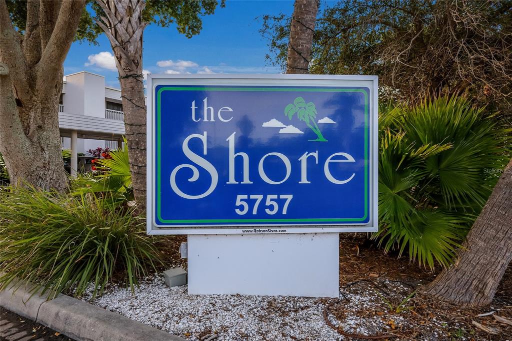A blue sign with white text reading The Shore 5757 stands outdoors near palm trees — 5757 GULF OF MEXICO DRIVE UNIT 205, LONGBOAT KEY, FL