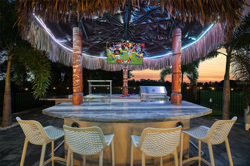 An outdoor tiki bar with a stone countertop, four white chairs, and a mounted television — 801 GOLDEN BEAR DRIVE, KISSIMMEE, FL