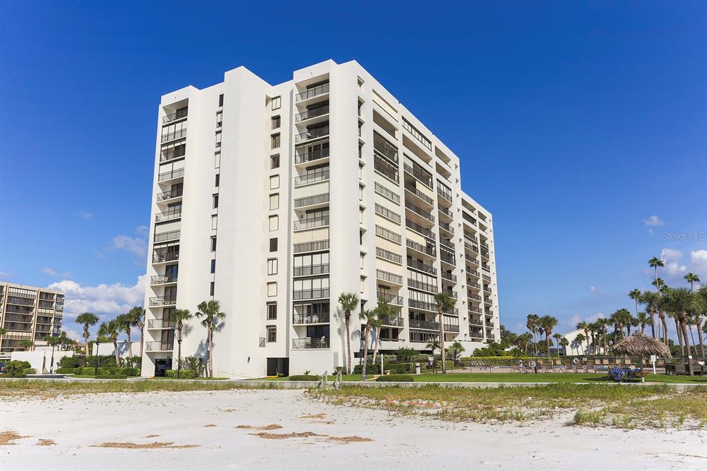 A tall, white multi-story residential building stands behind a sandy beach under a blue sky — 1460 GULF BLVD UNIT 412, CLEARWATER BEACH, FL