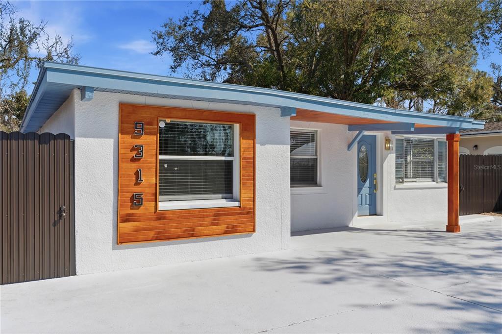 A white house exterior features a wood-paneled window accent, a blue front door, and driveway — 9315 N 16TH STREET, TAMPA, FL