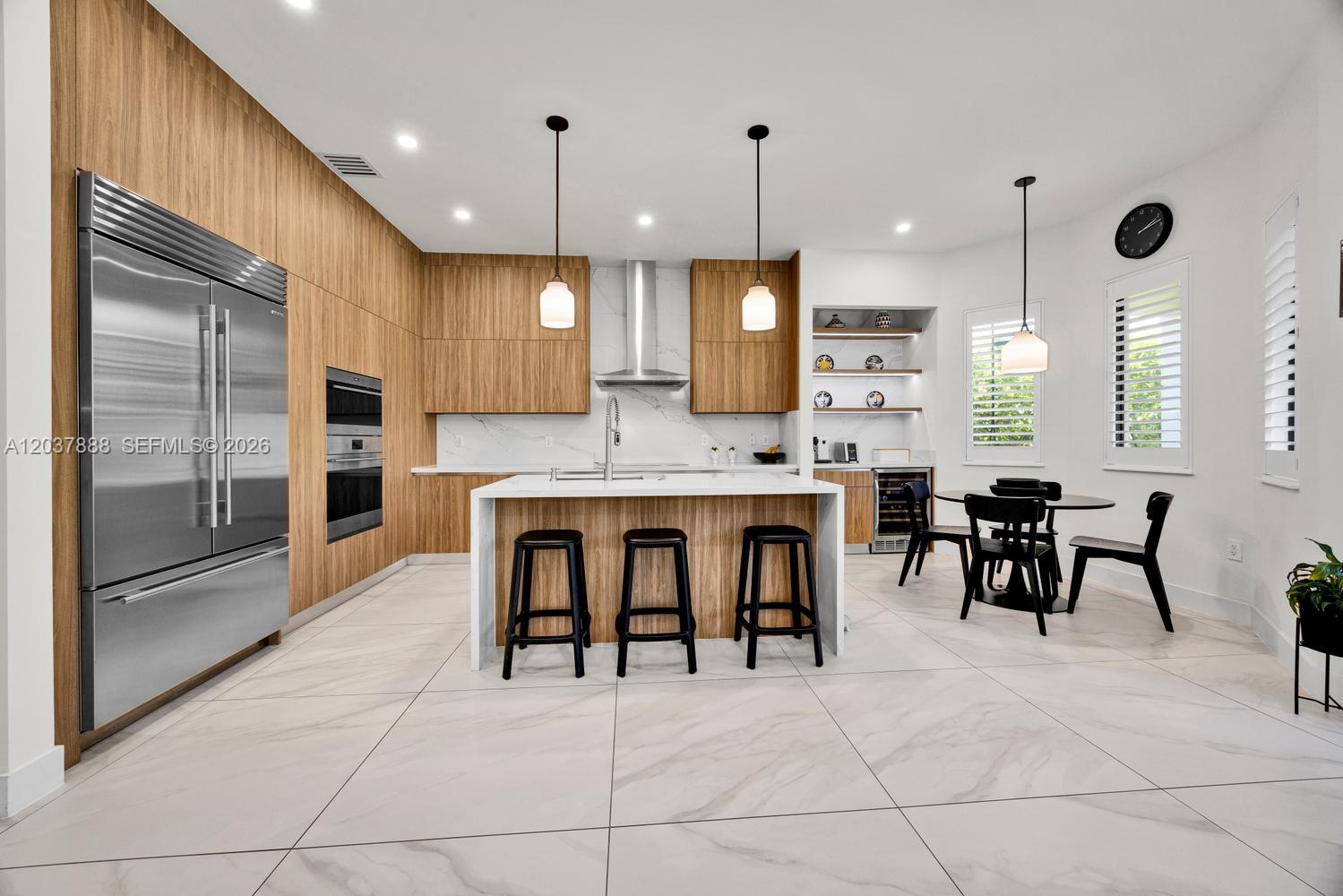 A modern kitchen features wood cabinetry, a white marble island, and a dining area — 12021 SW 82nd Pl, Miami FL 33156, MIAMI, FL