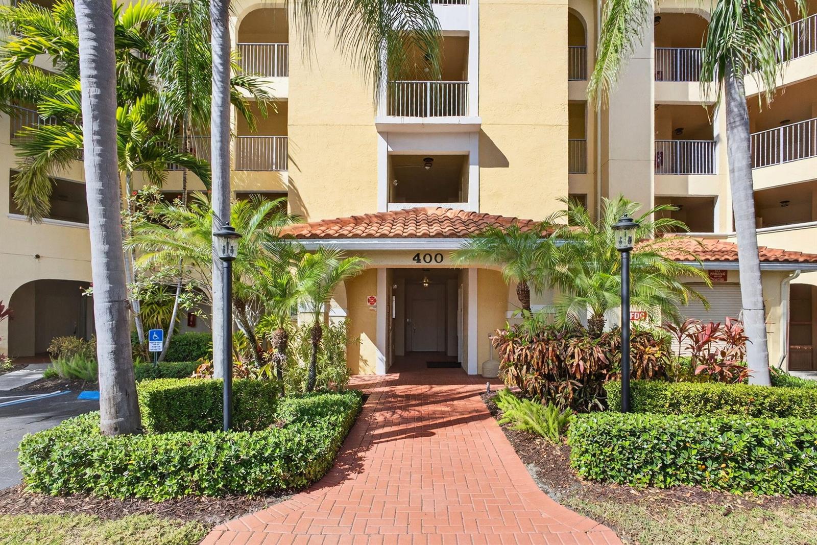 A brick walkway leads to the entrance of a multi-story yellow apartment building — 400 Uno Lago Drive # 103, Juno Beach FL 33408, JUNO BEACH, FL